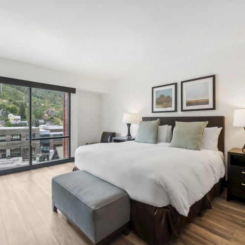 A modern bedroom with a large bed, two lamps, framed art, and a view of a hillside through a sliding glass door.