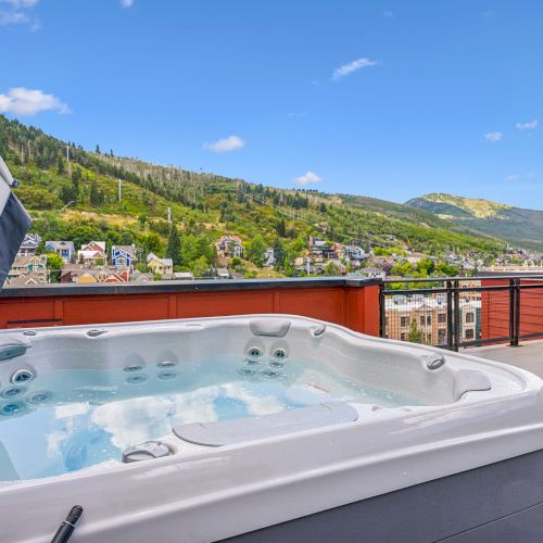 A rooftop hot tub overlooks a scenic hillside with houses and lush greenery under a clear blue sky.