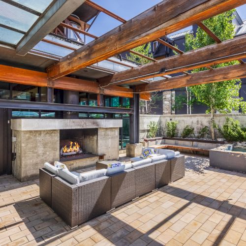 The image shows a modern outdoor patio with a seating area, fireplace, and wooden pergola, surrounded by a garden.