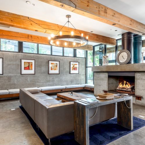 A modern living room with a concrete fireplace, leather sofas, framed art, and wooden beams under ceiling lights, creating a cozy ambiance.