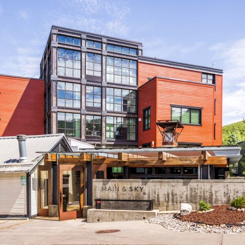 The image shows a modern building with red and black accents, large windows, and a green tree beside it, under a clear blue sky.