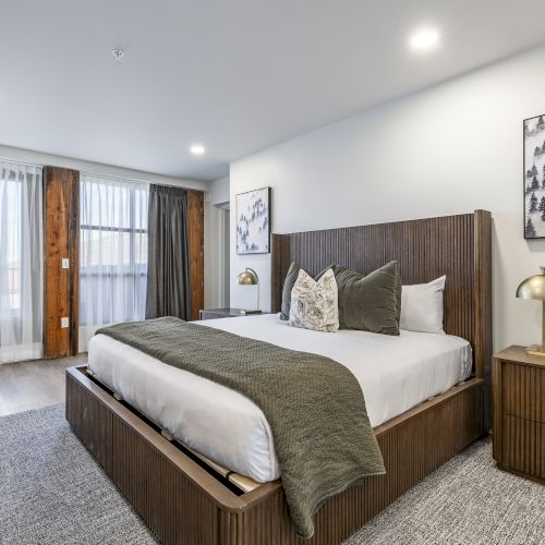 A modern bedroom with a wooden bed, gray bedding, wall art of snowy trees, and lamps plus curtains by the window.