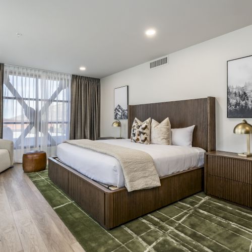 A modern bedroom with a large bed, brown curtains, framed mountain art, and gold bedside lamps, styled with green runner rugs.