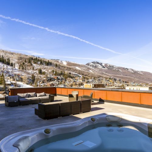 A sunny snowy mountain town view from a deck with outdoor seating and a hot tub, framed by pine trees and blue sky.