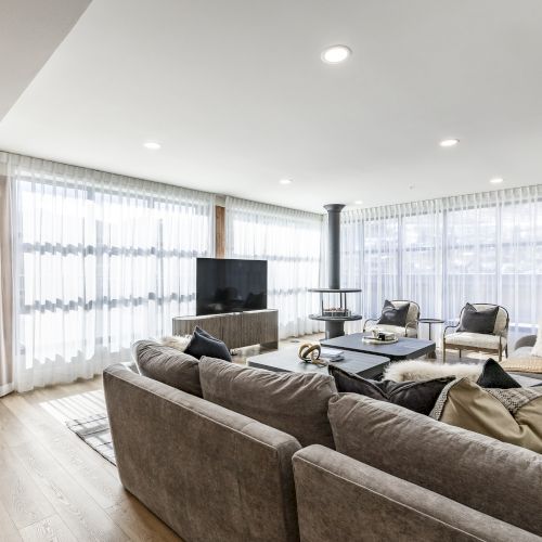 Bright modern living room with a gray sectional, TV on a cabinet, lamps, and large sheer curtains.