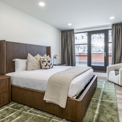 A modern bedroom with a large wooden bed, bedside lamp, window doors, and gray curtains, overlooking a neighborhood view.