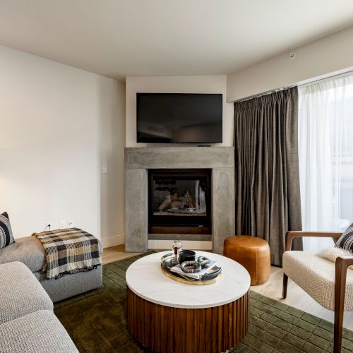 A cozy living room with a mounted TV above a fireplace, upholstered seating, and a round coffee table by large windows.