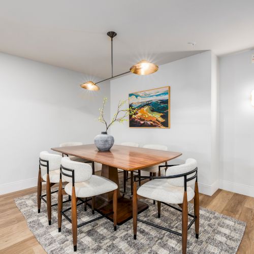 Bright dining room with a wooden table, white upholstered chairs, pendant lights, wall art, and a potted plant.