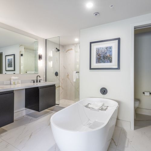 The image shows a modern bathroom with a freestanding bathtub, double vanity, wall mirrors, and a separate toilet area, featuring sleek decor.