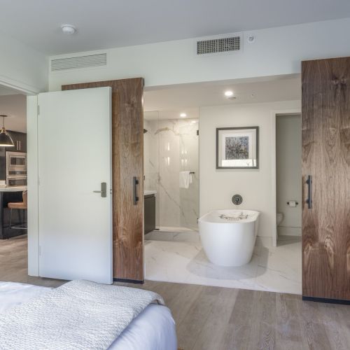 The image shows a modern bedroom with an open doorway leading to a bathroom featuring a freestanding bathtub and wooden sliding doors.