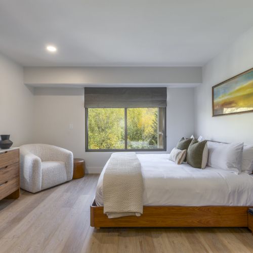 A modern bedroom with wooden furniture, a large bed, a dresser, an armchair, and a window with a scenic view and a painting on the wall.