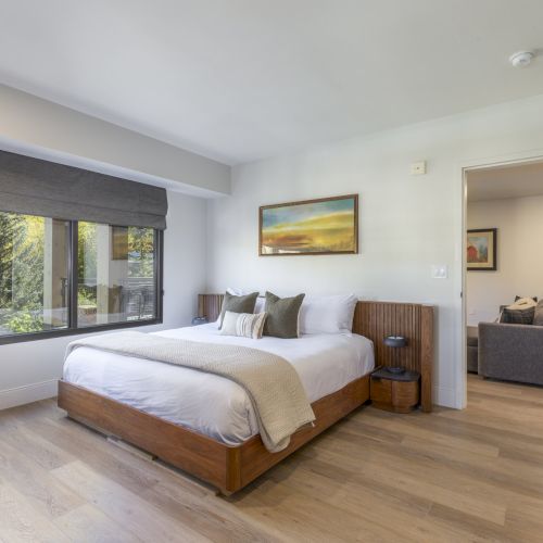 A modern bedroom with a large bed, window with blinds, and a painting on the wall, connected to a sitting area through an open doorway.