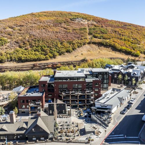 Aerial view of a small town with modern buildings, surrounded by a hill with autumn foliage under a clear blue sky.