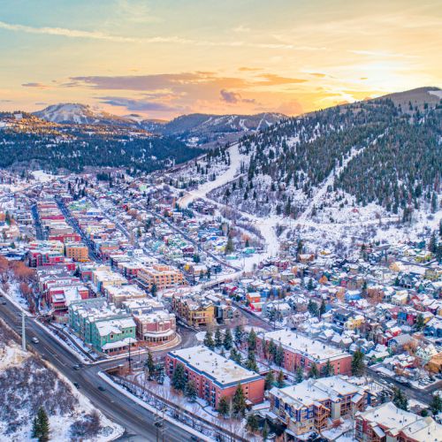 Aerial view of a snowy mountain town at sunset, with winding roads, buildings, and pine forests on snowy slopes.