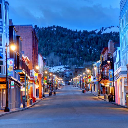 A lit winter street with shops, colorful banners, lamp posts, and snow-covered hills in the distance under a blue dusk sky.