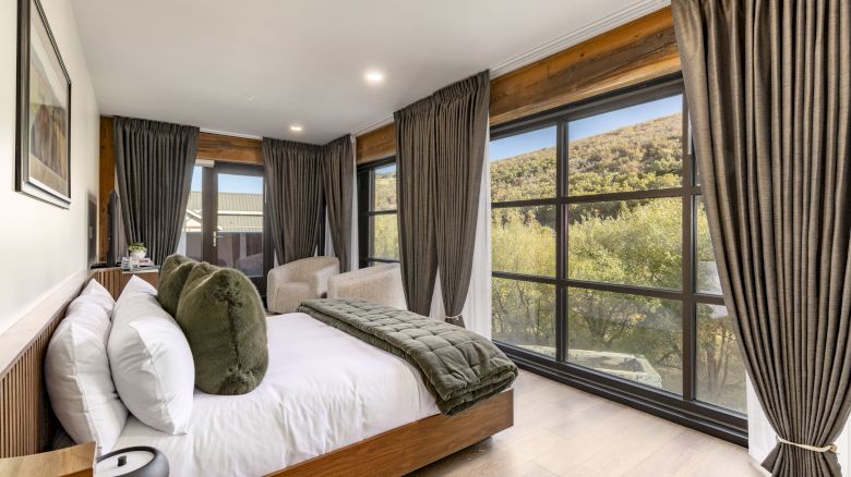 A cozy bedroom with a double bed, green pillows, large windows with curtains, and a scenic outdoor view.