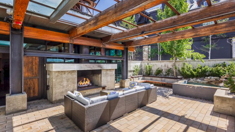 The image shows a modern outdoor patio with a fireplace, seating area, wooden beams, and greenery in a landscaped setting.