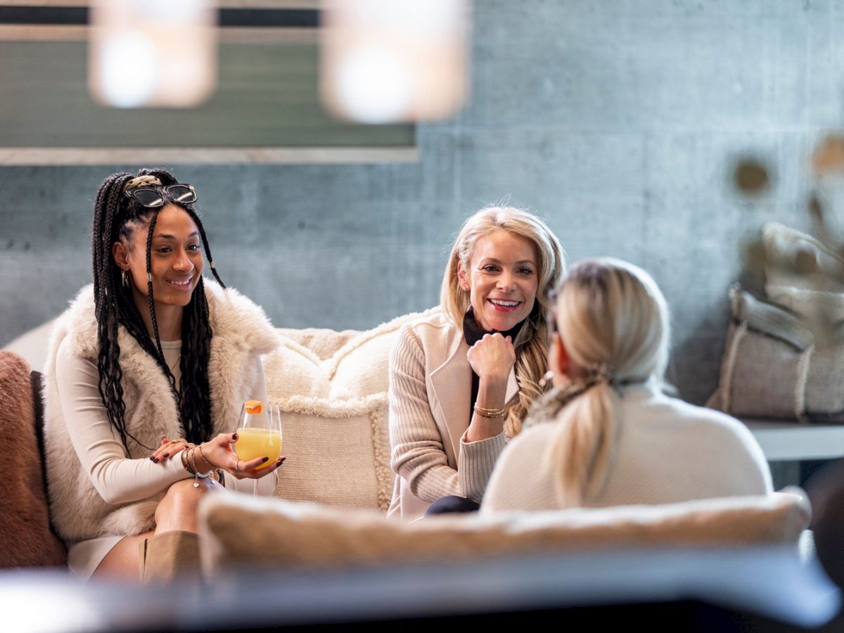 Three women in cozy sweaters chat on a couch in bright living room, smiling while one holds a glass of orange drink.