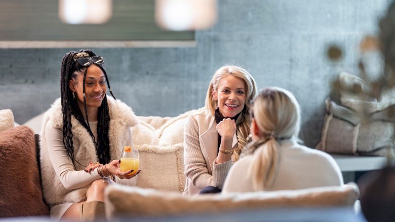 Three women in cozy sweaters chat on a couch in bright living room, smiling while one holds a glass of orange drink.