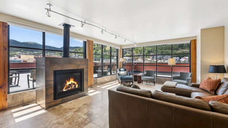 A modern living room with large windows, a central fireplace, leather sofas, and mountain views.