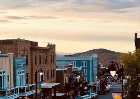 A quaint street scene with cars parked, buildings on either side, and hills in the distance under a colorful sky.