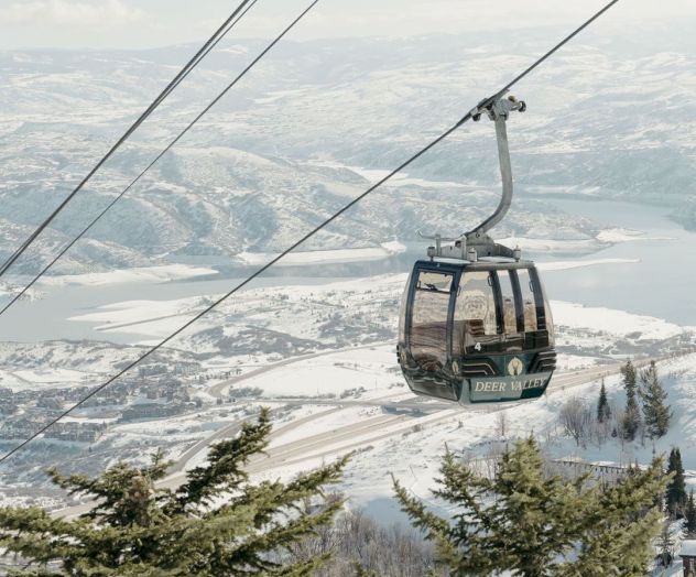 A ski lift gondola is suspended over a snowy landscape with trees and distant hills in the background.