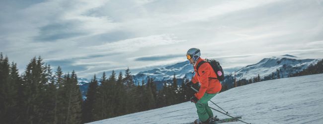 A skier in a bright jacket is skiing down a snowy slope with mountains and pine trees in the background, under a partly cloudy sky.