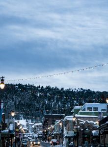 A snowy, lantern-lit street lined with buildings, leading to a tree-covered hill under a cloudy sky, with twinkling string lights above.