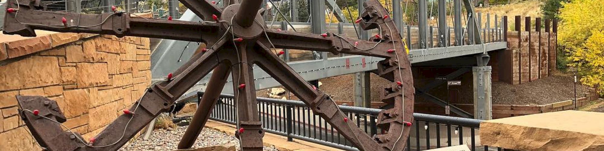 The image shows a large, rusted gear-like structure on display outdoors, with an informative plaque and a bridge in the background.