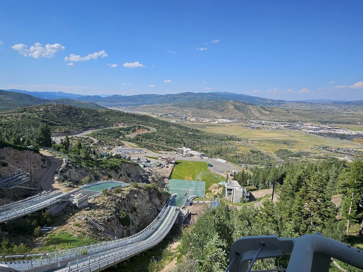 A scenic view of a landscape with a ski jumping facility, surrounded by hills, trees, and a wide valley under a blue sky with clouds.