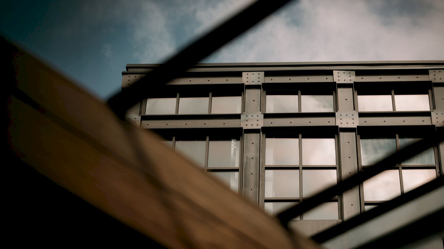 A modern building facade with steel beams and grid-like window panels, seen through dark structural bars against a cloudy sky.