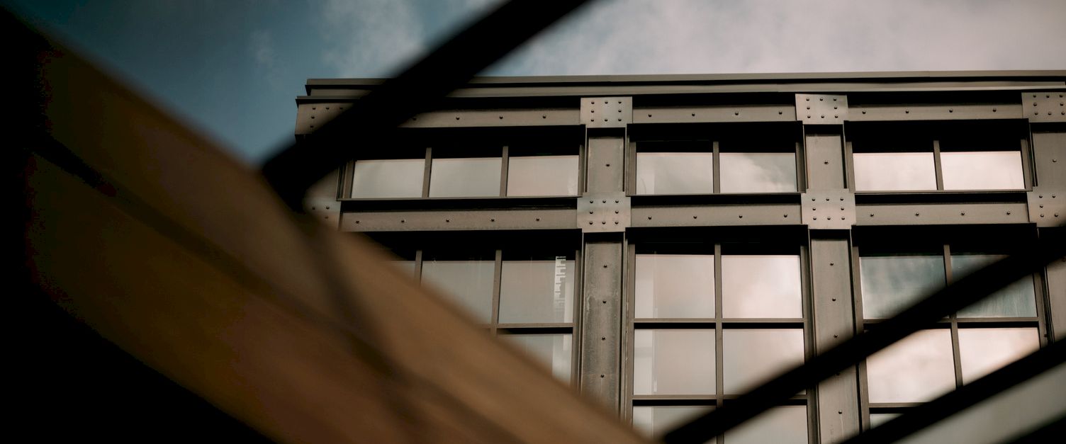 A modern building facade with steel beams and grid-like window panels, seen through dark structural bars against a cloudy sky.