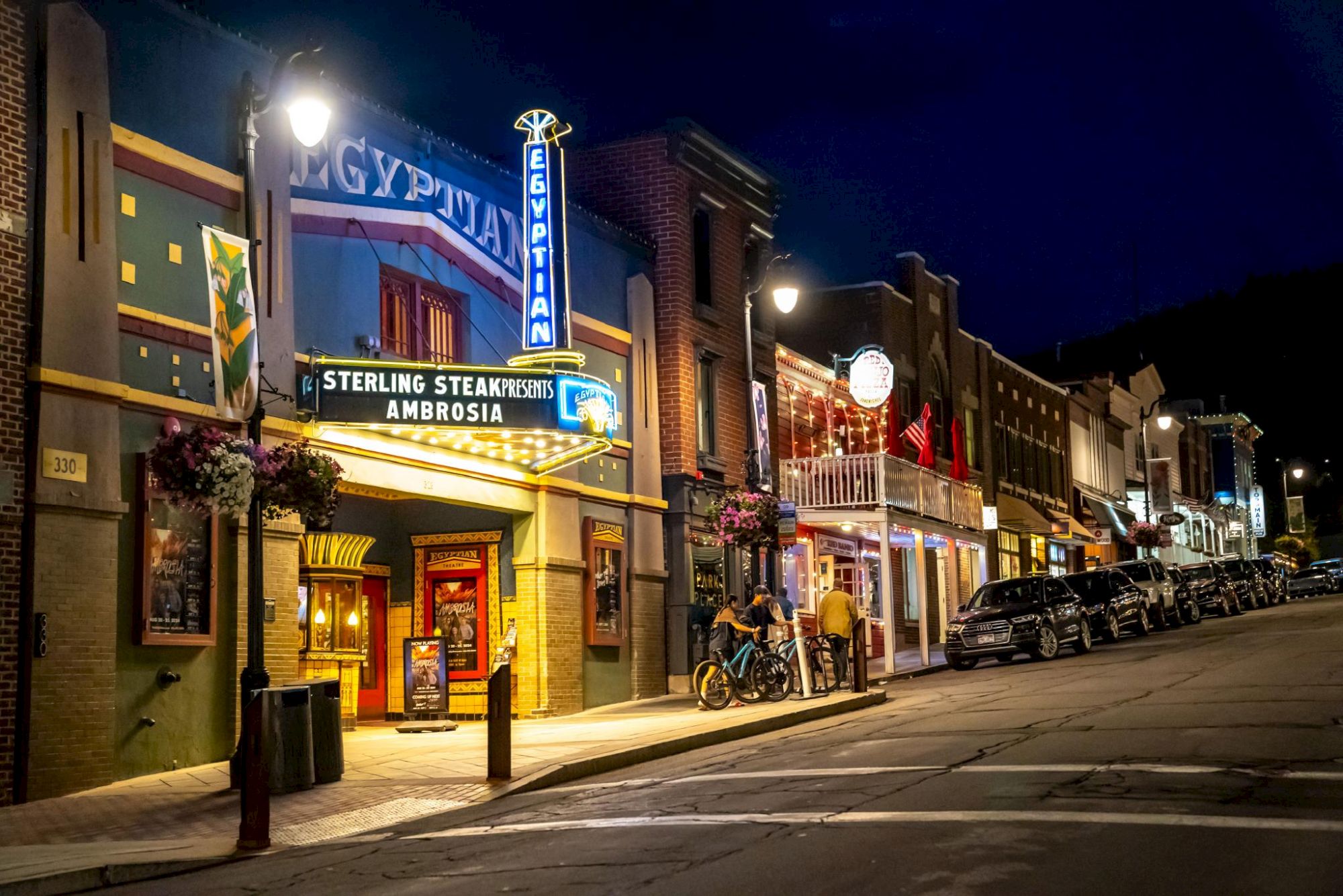A nighttime street in a small town, neon signs, a row of brick storefronts with diners and shops, quiet and lit by streetlights.