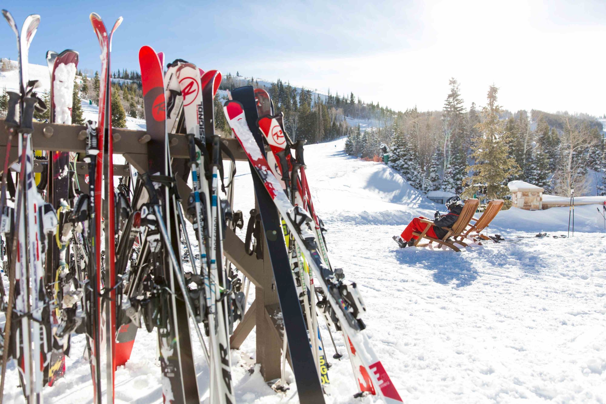 Ski gear leans against a rack with snow-covered slopes and a bench in the bright, sunny winter scene.