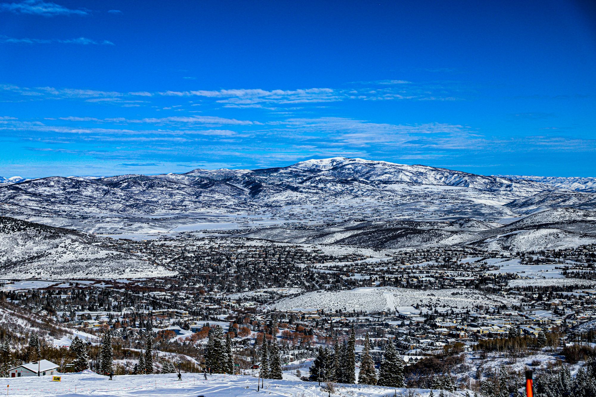 A snowy mountainous landscape with a clear blue sky, small town below, and trees scattered across the snowy terrain.