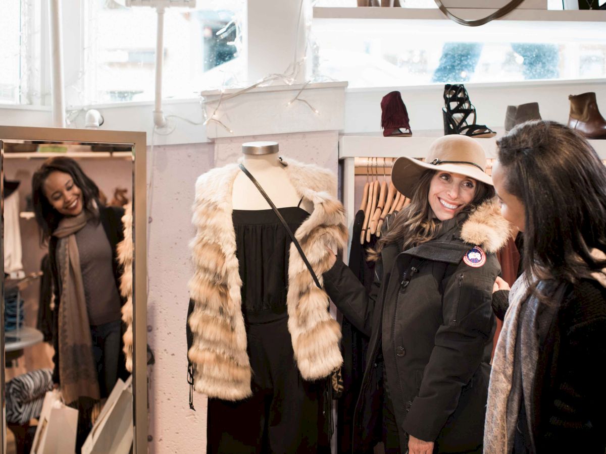 Three women shop in a fitting room area with coats and a fur jacket; one tries on a hat while another checks a mirror.
