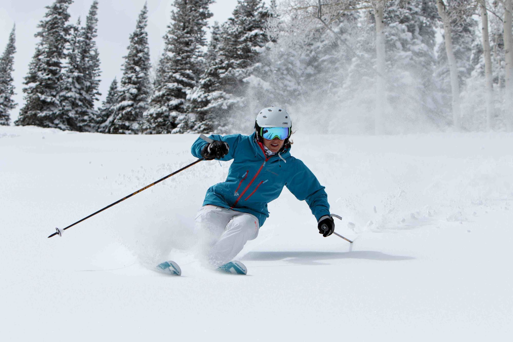 A skier in a blue jacket and white pants carves through fresh powder, helmeted and gliding hard, sending a spray of snow to the side.