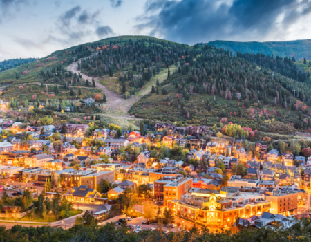 A small town nestled in a valley with warm streetlights, surrounded by forested hills and rolling green slopes under a partly cloudy sky.