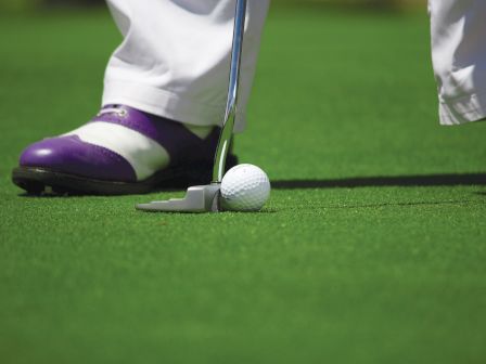 A close-up of a golfer lining up a putt: golf ball near the hole, club face ready, purple-and-white shoes on a green putting surface.