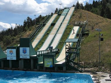 A large outdoor swimming pool with a tall multi-lane water slide on a grassy hill, blue sky, and a few clouds in the background.