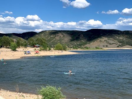 A sunny lakeside beach with swimmers in the water and hills in the background under a partly cloudy sky.