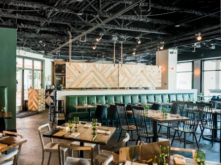 A modern restaurant interior with wooden tables, green upholstered booths, chic chairs, and a long bar area under bright overhead lighting.