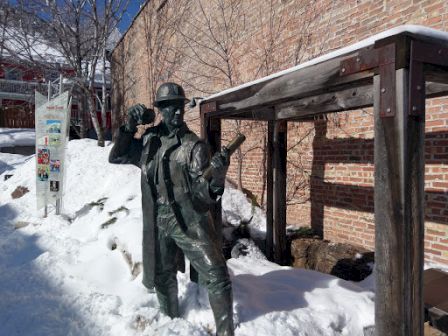 A bronze statue of a soldier in snow beside a brick building, saluting with one hand and holding a rifle in the other.
