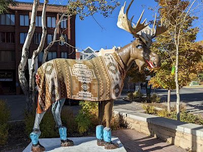 A statue of a deer wearing a patterned poncho and blue boot covers, standing in a campus-like plaza with autumn trees in the background.