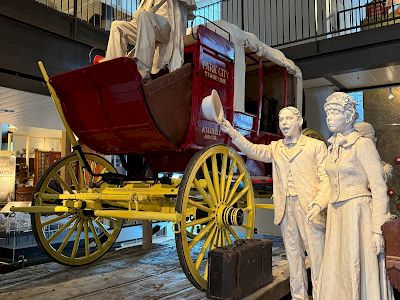 A red antique carriage with yellow wheels and statues of a driver and two passengers beside it, indoors in a museum.