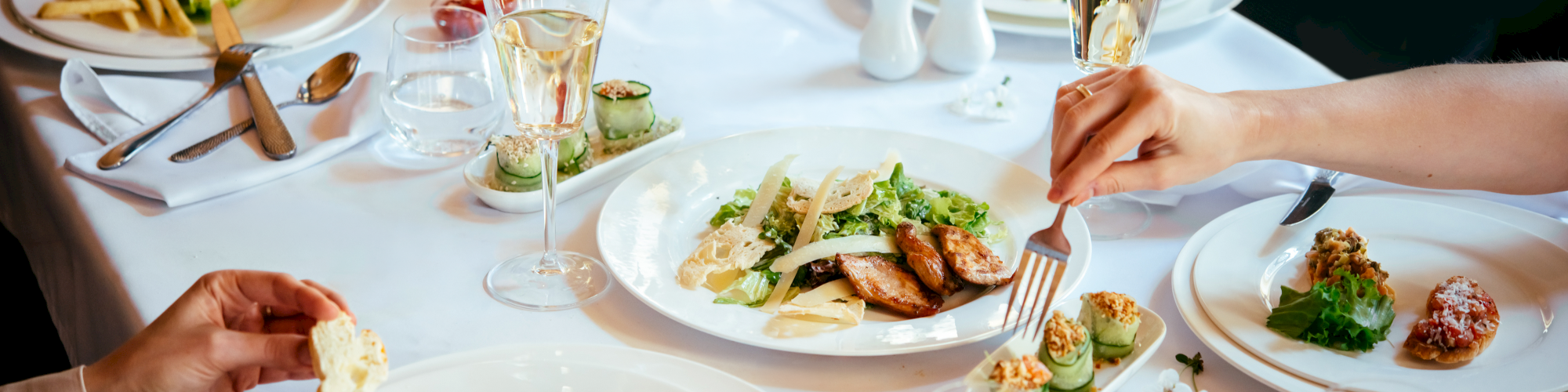 A group sits around a table enjoying a shared meal with plated dishes, wine glasses, and cutlery, as they toast and dine together.