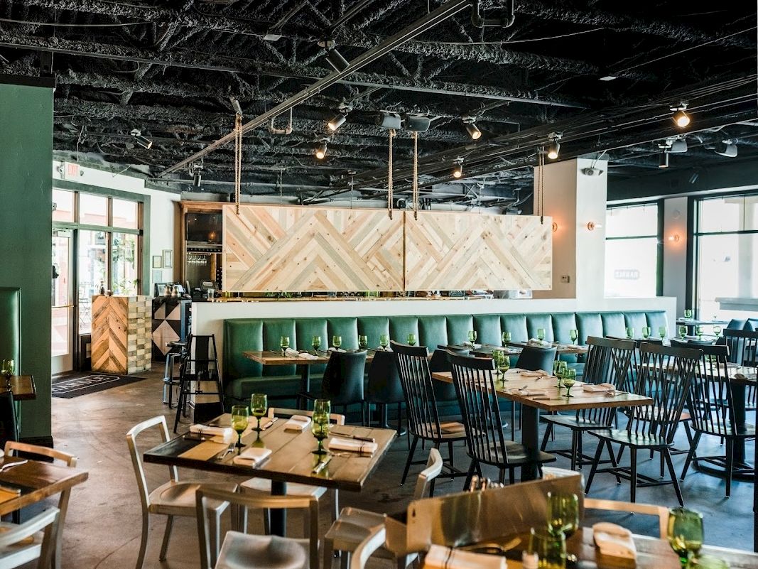A modern restaurant interior with wooden tables, mixed chairs, green upholstered bar seating, pendant lights, and natural light from large windows.