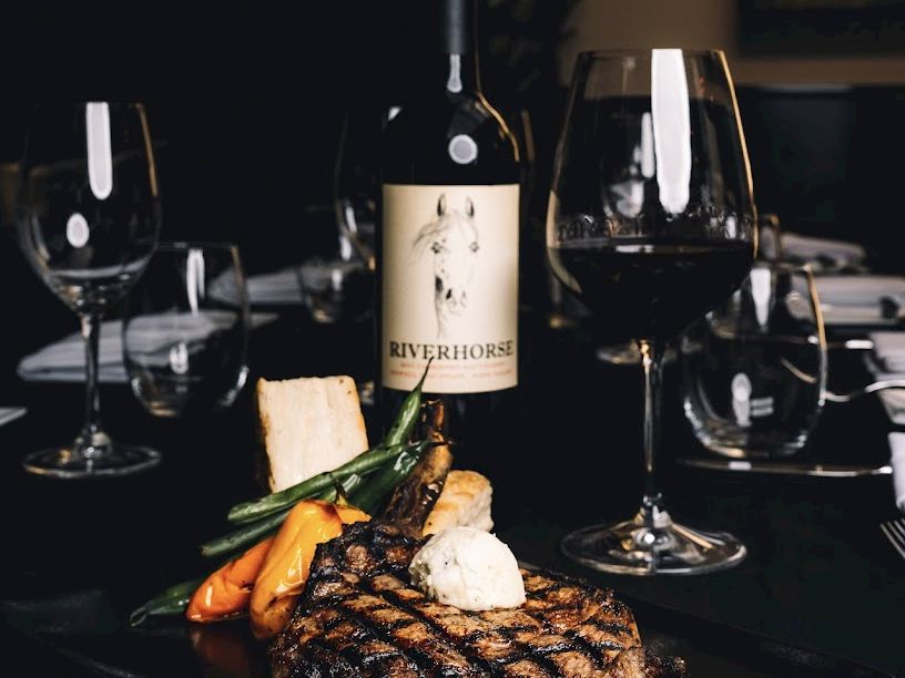 A fine dining scene with a steak, vegetables, a dollop of sauce, wine bottle, and glasses on a dark table.