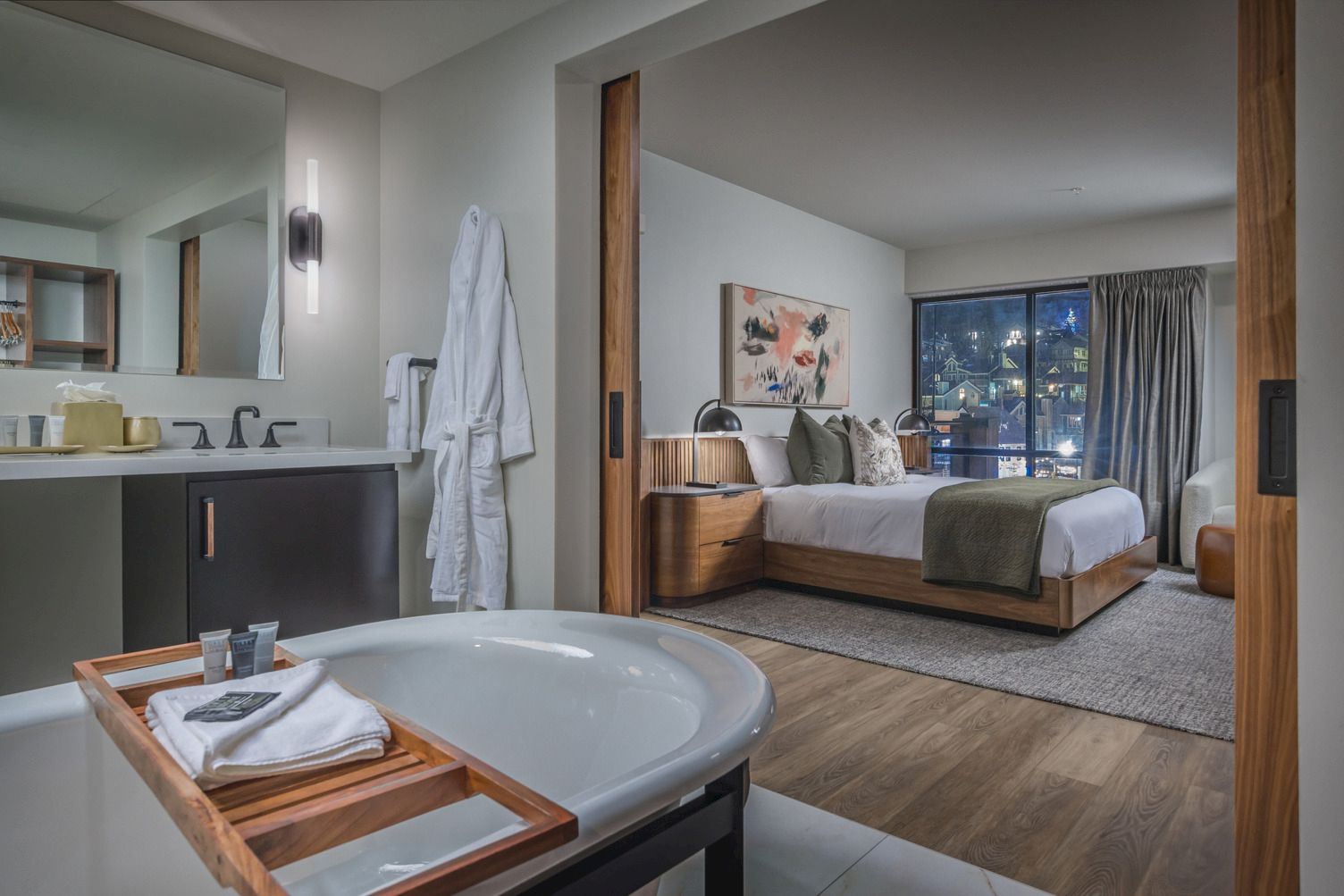 Modern hotel suite with a bathtub and toiletries in the foreground and a cozy bedroom with a bed and city night view.