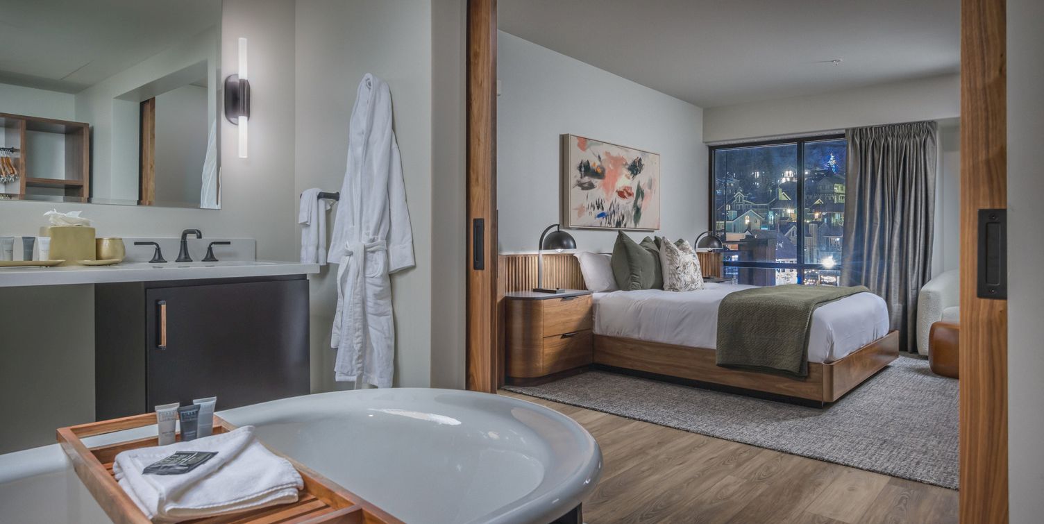 Modern hotel suite with a bathtub and toiletries in the foreground and a cozy bedroom with a bed and city night view.